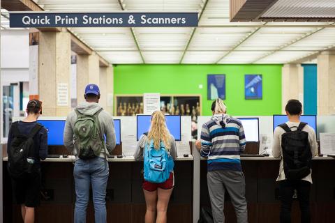 row of students, back to camera, looking at computer kiosks