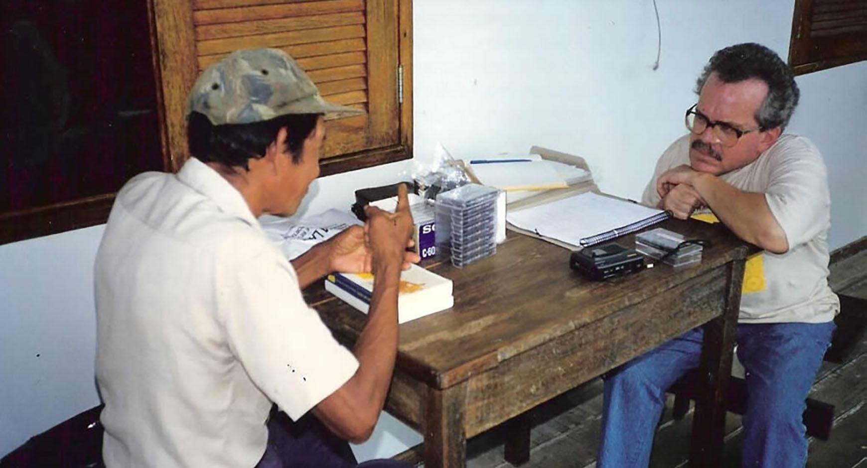 two men having a discussion at a wooden table with audio cassettes and notebooks on it.