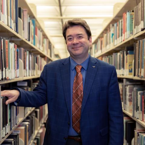 Robert H. McDonald, Senior Vice Provost and Director of the University of Texas Libraries, standing in an aisle of the Perry-Castañeda Library stacks wearing a blue checkered suit and burnt orange tie, smiling slightly with his hand resting on a bookshelf.