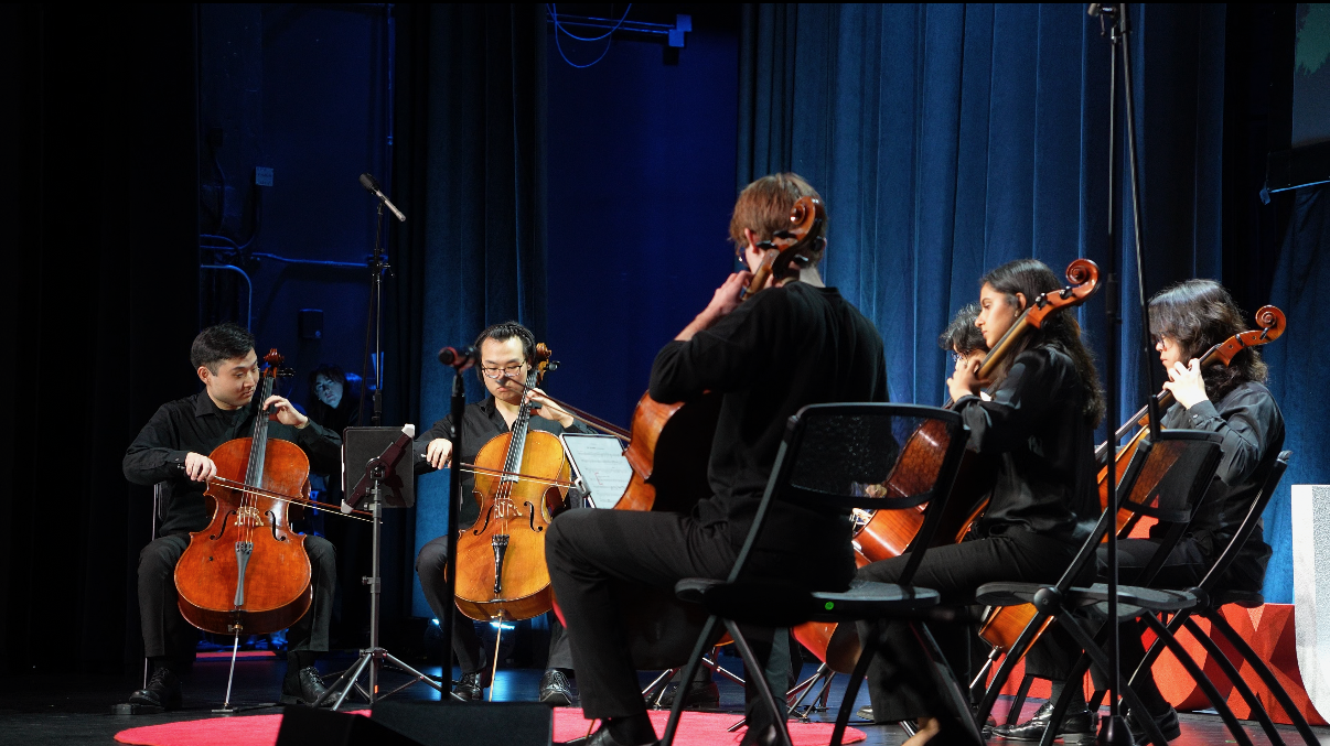 Photograph of a small cello ensemble performing on a stage with blue curtains. Several musicians sit in a semicircle, each playing a cello and reading from music stands, while microphone stands and cables are visible around them. The cellos are wooden with warm brown tones, and the performers wear dark concert attire under stage lighting.