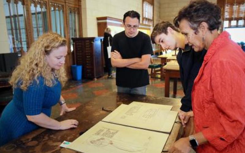 Image is a photo of the Architecture & Planning Library Special Collections area. Four people are stood around a table admiring an architectural rendering.