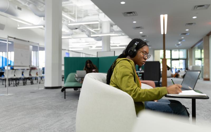 A photo of a student sitting in a comfy chair with headphones on writing something in a notebook