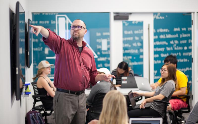 An instructor in a button down shirt pointing at a monitor standing front of a students